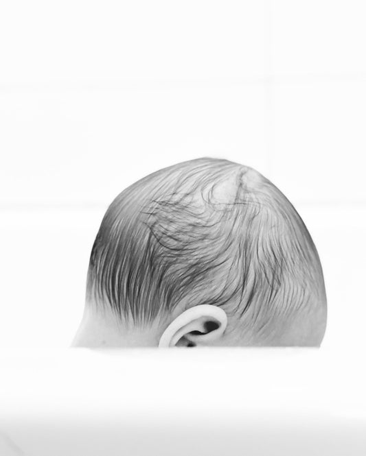 Black and white fine art photography print of a baby's soft hair from behind in tender intimate close-up against bright white background, by Israeli artist Tamar Green