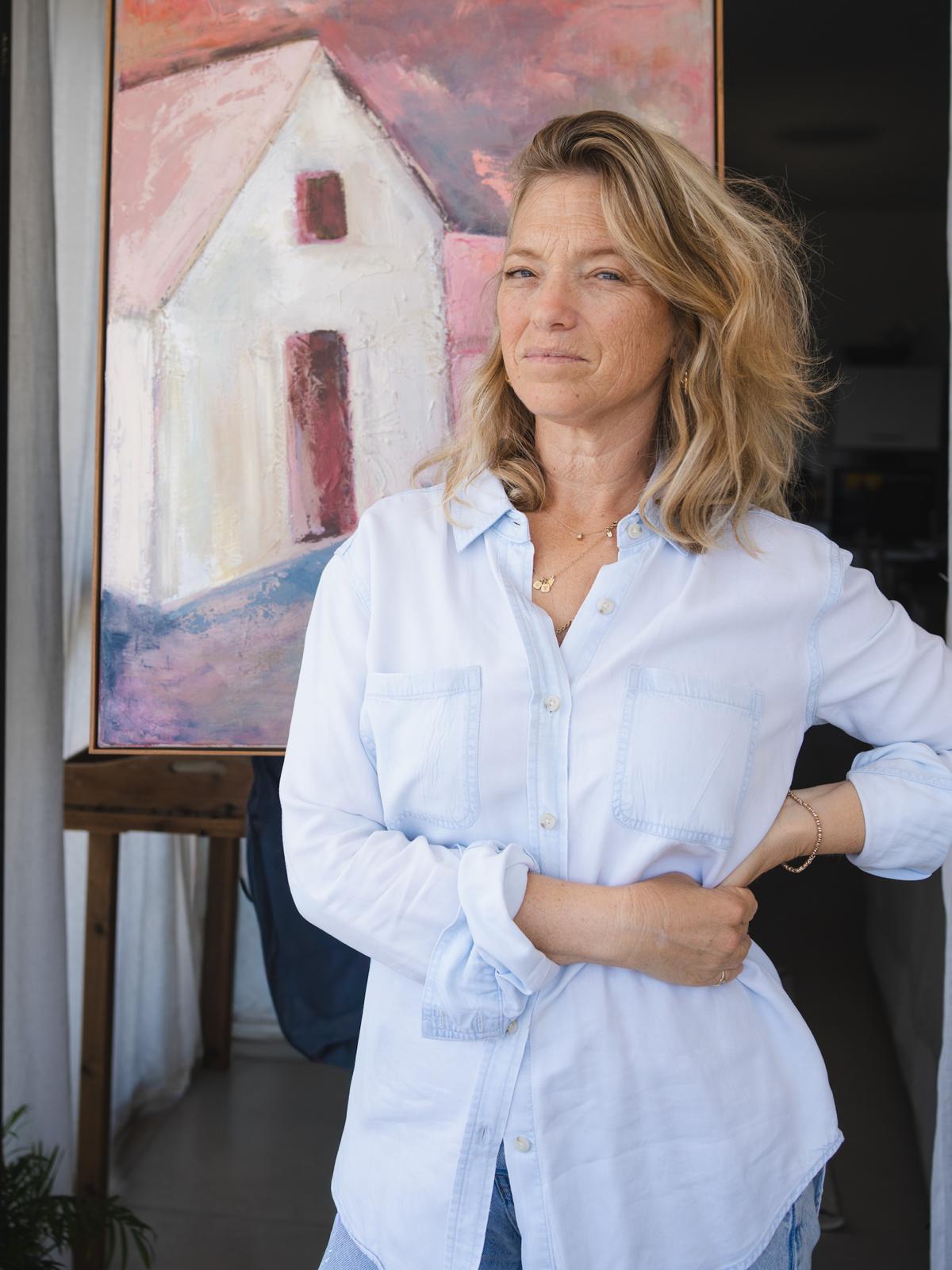 Portrait of Israeli artist Tamar Green in her studio, standing in front of a large oil painting of a white house under a pink sky from the Home series.