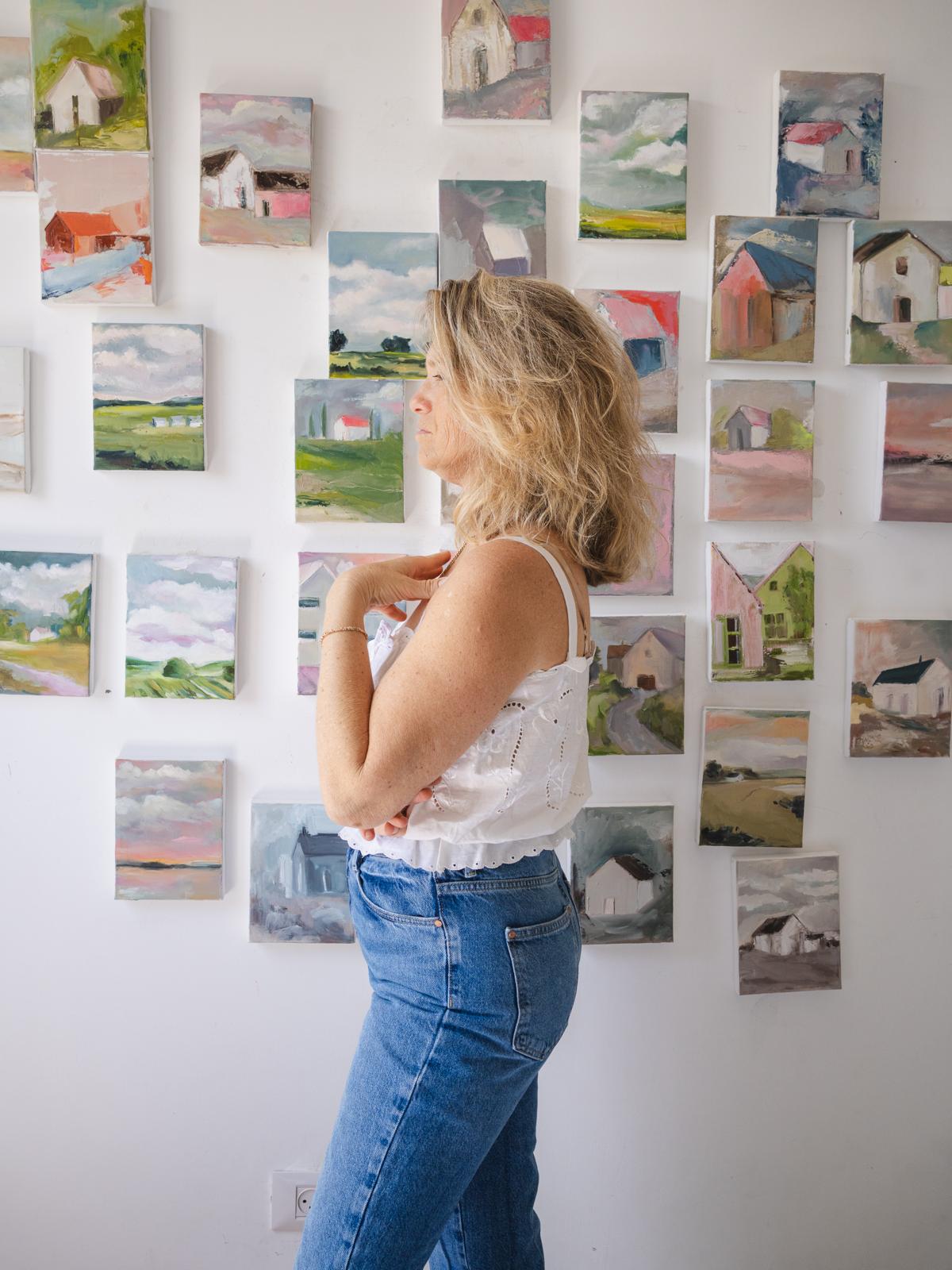 Israeli artist Tamar Green in her studio beside a white wall hung with dozens of small oil paintings of houses and landscapes from the Home series.